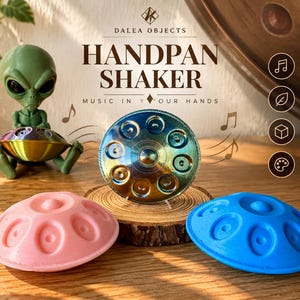 Könnte beinhalten: Drei Handpan-Shaker in Pink, Blau und irisierendem Metallic-Finish. Der Text lautet DALEA OBJECTS HANDPAN SHAKER MUSIC IN YOUR HANDS. Zu den Merkmalen gehören ein klarer Perkussionsklang, leicht und tragbar, 3D-gedruckt und in mehreren Farben erhältlich.