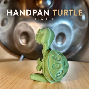 Könnte beinhalten: Eine hellgrüne Schildkrötenfigur mit einem Panzer, der kreisförmige Vertiefungen aufweist, vor einem metallischen Handpan. Der Text HANDPAN TURTLE FIGURE ist in weißen und beigen Buchstaben über der grünen Schildkrötenfigur zu sehen.