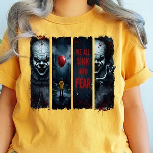May include: Mustard yellow t-shirt with a horror-themed graphic. The design features a menacing clown face on each side, with the text "WE ALL SINK INTO FEAR" in the centre. A red balloon and a figure in a yellow raincoat are also depicted.