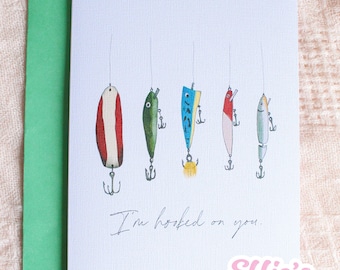 May include: Greeting card with a green envelope featuring a watercolor illustration of five colorful fishing lures hanging from strings. The text "I'm hooked on you" is handwritten below the lures. The Effie's Paperie logo is in the bottom right corner.