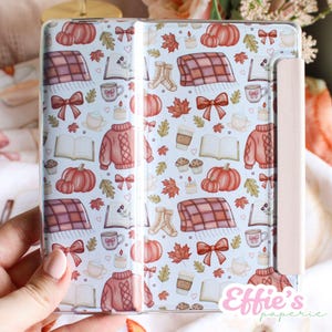 May include: A clear case with a pale pink border, decorated with a fall-themed pattern. The design includes pumpkins, sweaters, leaves, books, coffee cups, and bows in orange, red, and brown on a light blue backdrop.