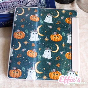 Könnte beinhalten: Eine tealfarbene Tablethülle mit einem Halloween-Muster. Das Design umfasst lächelnde orangefarbene Kürbisse, weiße Geister mit Blumenkränzen, Halbmonde, Fledermäuse und Sterne. Die Hülle hat einen hellrosa Magnetverschluss und den Text "Effie's paperie".