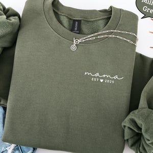 Könnte beinhalten: Militärgrünes Sweatshirt mit dem Wort "mama" und "EST 2025" in Weiß aufgedruckt. Eine silberne Halskette mit Sonnenanhänger liegt über dem Sweatshirt. Das Sweatshirt hat einen Rundhalsausschnitt und lange Ärmel.