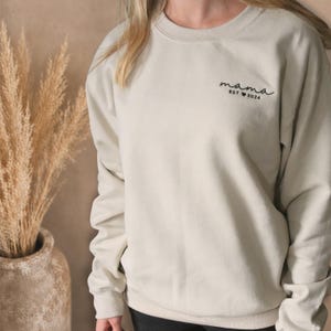 Pode incluir: Camisola bege com a palavra "mama" em escrita cursiva e "EST 2024" impressa por baixo. A camisola é de cor neutra e tem gola redonda. As mangas são compridas e o material parece macio.