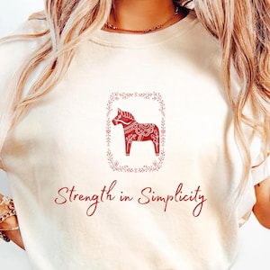 May include: Cream-colored t-shirt featuring a red Dala horse design within a floral frame. The phrase "Strength in Simplicity" is written in red script below the graphic. The shirt is paired with blue jeans.