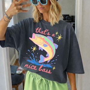 K&ouml;nnte beinhalten: Dunkelgraues T-Shirt mit einem bunten Fisch-Grafik und dem Text "that's a nice bass". Das Shirt wird mit hellgr&uuml;nen Shorts kombiniert. Das Bild wird vor einem Spiegel aufgenommen.