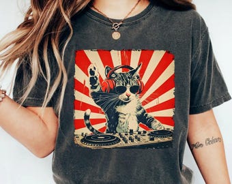 Op de afbeelding: Donkergrijs t-shirt met een kat-DJ-afbeelding. De kat draagt een zonnebril en een koptelefoon, met een rood-witte zonnestraal achtergrond. De kat staat achter een DJ-mixer, met een poot omhoog.