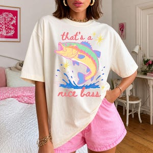 K&ouml;nnte beinhalten: Cremefarbenes T-Shirt mit einer farbenfrohen Illustration eines springenden Bassfisches und dem Text "that's a nice bass". Der Fisch ist in Gelb-, Gr&uuml;n-, Blau- und Rosat&ouml;nen dargestellt. Das Shirt wird mit rosa Shorts getragen.
