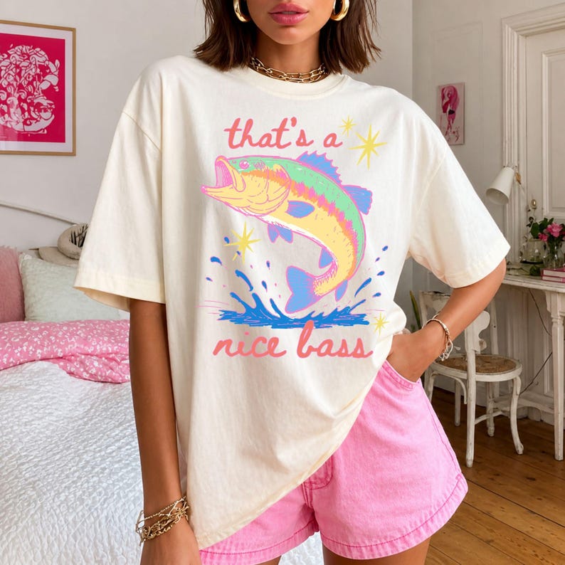 K&ouml;nnte beinhalten: Cremefarbenes T-Shirt mit einer farbenfrohen Illustration eines springenden Bassfisches und dem Text "that's a nice bass". Der Fisch ist in Gelb-, Gr&uuml;n-, Blau- und Rosat&ouml;nen dargestellt. Das Shirt wird mit rosa Shorts getragen.