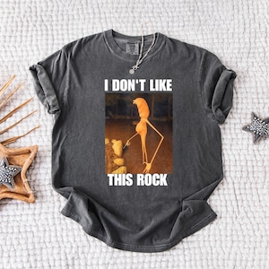 May include: Dark gray t-shirt with the text "I DON'T LIKE THIS ROCK" above an image of a stick figure pointing at a rock formation. The shirt has a crew neck and short sleeves.