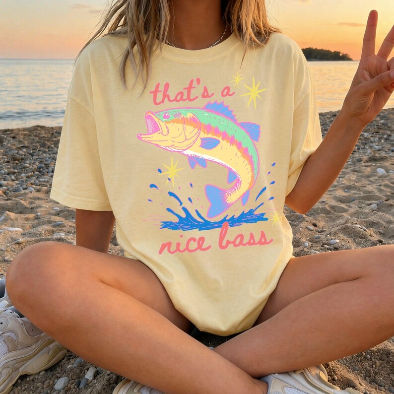 K&ouml;nnte beinhalten: Hellgelbes T-Shirt mit einer farbenfrohen Grafik eines springenden Bassfisches. Der Text "that's a nice bass" ist in Rosa und Blau &uuml;ber und unter dem Fisch aufgedruckt. Das Shirt wird am Strand getragen.