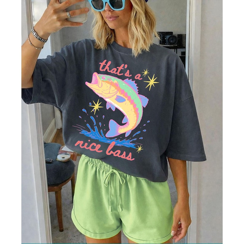 K&ouml;nnte beinhalten: Dunkelgraues T-Shirt mit einem bunten Fisch-Grafik und dem Text "that's a nice bass". Das Shirt wird mit hellgr&uuml;nen Shorts kombiniert. Das Bild wird vor einem Spiegel aufgenommen.