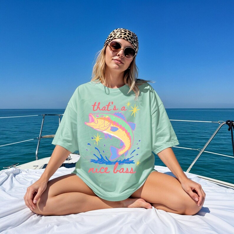 K&ouml;nnte beinhalten: Hellgr&uuml;nes T-Shirt mit dem Text "that's a nice bass" und einer bunten Fischgrafik. Das Bild zeigt das Shirt auf einem Boot vor blauem Himmel und Wasser.