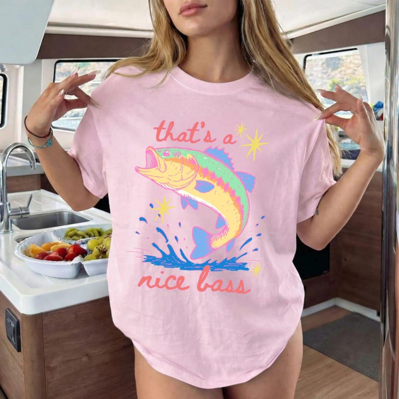 K&ouml;nnte beinhalten: Hellrosa T-Shirt mit einer farbenfrohen Illustration eines springenden Bassfisches. Der Text "that's a nice bass" ist oberhalb und unterhalb des Fisches in einer verspielten Schriftart aufgedruckt. Das Shirt hat kurze &Auml;rmel.