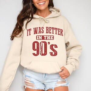 May include: A beige hooded sweatshirt featuring the text IT WAS BETTER IN THE 90'S printed in a distressed red font across the chest. The hoodie includes a drawstring hood, long sleeves, and a front kangaroo pocket for a casual look.