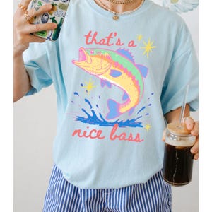 K&ouml;nnte beinhalten: Hellblaues T-Shirt mit einem farbenfrohen Motiv eines springenden Fisches und dem Text "that's a nice bass". Der Fisch ist gelb, rosa, gr&uuml;n und blau. Der Text ist rosa. Ein l&auml;ssiges T-Shirt.