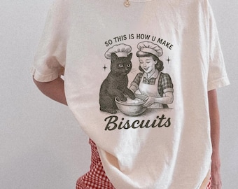 May include: Cream-colored t-shirt featuring a vintage-style illustration of a black cat and a woman in a chef's hat and apron, mixing ingredients in a bowl. The text "SO THIS IS HOW U MAKE Biscuits" is printed below the illustration.