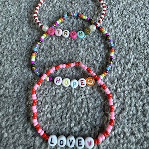 May include: Three beaded bracelets with positive messages. The top bracelet is red and white. The middle bracelet has the word "STRONG" spelled out in colourful letter beads. The bottom bracelet has the words "HOPE" and "LOVE" spelled out in letter beads.