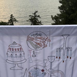 以下が含まれることがあります： ケーキ、ロブスター、パスタ、ワイングラス、シャンデリアの刺繍イラストが施された白いテーブルクロス。デザインは茶色、赤、青の色合いです。背景には海の景色が広がっています。