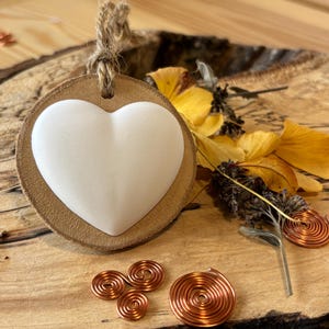 May include: A white heart-shaped stone sits on a round, light brown leather pendant with a jute cord. Copper wire spirals and dried yellow flowers are arranged on a wooden surface, creating a natural, earthy aesthetic.