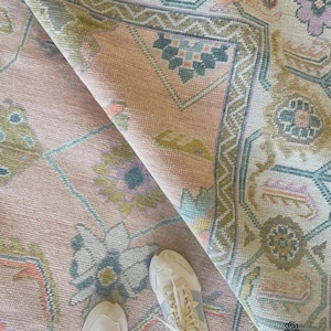 Pode incluir: Um tapete dobrado, em estilo vintage, em tons de rosa, verde e azul. O tapete apresenta um padrão floral e geométrico detalhado com uma aparência desgastada. O canto do tapete é levantado, revelando a parte inferior. Tênis brancos são visíveis.