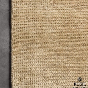 Puede incluir: Primer plano de una alfombra beige tejida con una superficie texturizada y un borde con flecos. La alfombra tiene un color natural y neutro y un patrón de tejido visible. El logotipo de Rosie Rug Store está en la esquina inferior derecha.