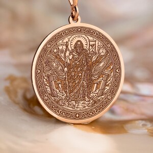 Puede incluir: Un collar con colgante de oro rosa con un medallón circular detallado con una figura religiosa grabada. El colgante cuelga de una delicada cadena. El collar se exhibe sobre una superficie nacarada.