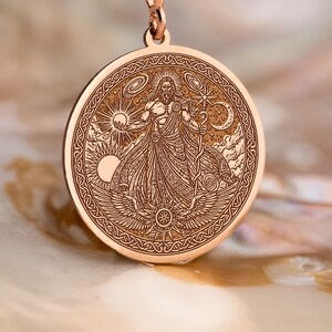 Könnte beinhalten: Halskette mit Anhänger aus Roségold. Der runde Anhänger zeigt ein eingraviertes Himmelsdesign, das eine Figur umgeben von Sonne, Mond und Sternenmotiven darstellt. Die Halskette wird auf einer perlmuttartigen Oberfläche präsentiert.