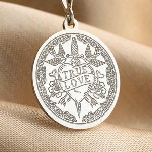 May include: Silver-toned pendant necklace with a round charm. The charm features a heart pierced by a dagger, the words "TRUE LOVE", doves, and floral accents. The necklace hangs on a delicate chain.