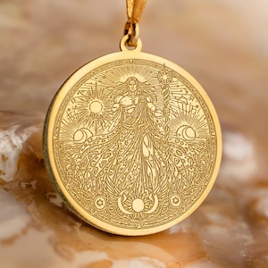 May include: A gold pendant necklace with an engraved design. The circular pendant shows a figure with sun and moon motifs. The necklace has a delicate chain and a simple clasp. The pendant is approximately 3.8 cm in diameter.