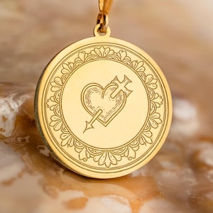 May include: Gold-colored pendant necklace featuring a circular charm with a heart pierced by an arrow and a cross. The charm is surrounded by a decorative border. The necklace is displayed on a textured surface.