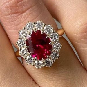 Peut inclure: Une bague en or ornée d'une grande pierre précieuse rouge foncé de taille ovale au centre, entourée d'un halo de douze pierres rondes, claires et facettées. La monture est en or jaune poli, présentant un design classique en grappe.
