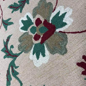 Könnte beinhalten: Beiger runder Teppich mit floralem Muster. Der Teppich zeigt eine große zentrale Blume mit weißen, grünen, braunen und burgunderfarbenen Blütenblättern. Grüne Blätter und burgunderfarbene Ranken sind über die Oberfläche des Teppichs verteilt. Der Teppich hat ein strukturiertes Aussehen.