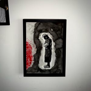 May include: Framed artwork featuring a black and white ink illustration of a person in a hat with arms crossed. The figure is set against a dark, abstract background with red accents. The artwork is encased in a black frame.