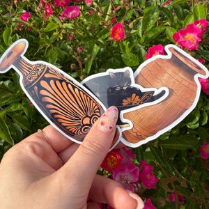 Peut inclure: Trois autocollants représentant des motifs de poterie grecque antique et un chat. Les autocollants sont tenus devant un fond de roses roses. Les autocollants de poterie sont marron et orange, et l'autocollant de chat est noir et gris.