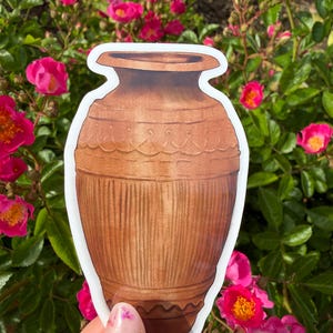 Peut inclure: Un autocollant représentant un vase marron avec une bordure blanche. Le vase présente un motif détaillé avec des lignes verticales et des bandes décoratives. L'autocollant est tenu devant un fond de roses roses et de feuillage vert.