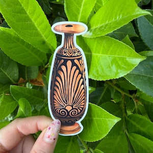 Peut inclure: Un autocollant représentant un vase orange et noir avec un dessus plat et des motifs complexes. Le vase présente un design symétrique avec une bordure blanche. L'autocollant est posé sur un fond de feuilles vertes.