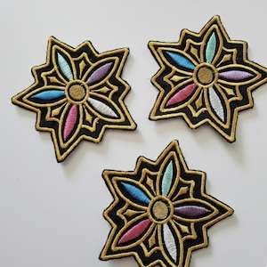 Op de afbeelding: Drie geborduurde patches in een stervormig ontwerp. Elke patch heeft een zwarte achtergrond, een gouden rand en kleurrijke bloemachtige elementen in roze, blauw, wit en paars. Het midden van elke patch is bruin.
