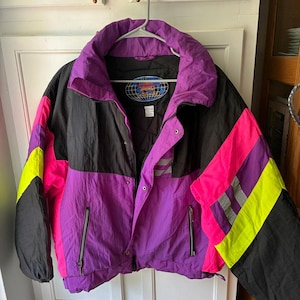 May include: A colour-blocked puffer jacket featuring a vibrant palette of purple, black, neon pink, and neon yellow. The jacket includes a high collar, a front zip closure with snap buttons, two zipped side pockets, and reflective silver stripes.