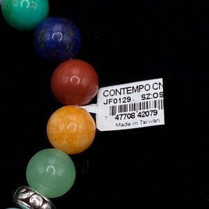 May include: A bracelet featuring a series of smooth, round, multi-colored gemstone beads in shades of turquoise, blue, red, orange, and light green, accented by a silver-toned metal spacer bead. A white rectangular tag with a barcode and text is attached.