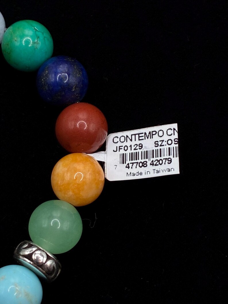 May include: A bracelet featuring a series of smooth, round, multi-colored gemstone beads in shades of turquoise, blue, red, orange, and light green, accented by a silver-toned metal spacer bead. A white rectangular tag with a barcode and text is attached.