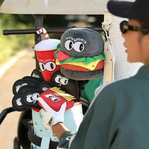 May include: Golf club head covers and accessories in a golf bag. The covers are designed as a hamburger, fries, and a soda cup, all with cartoon eyes. The golf bag is light blue and is in a golf cart.