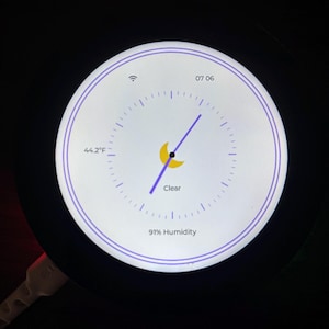 May include: A circular digital display shows the time, weather, and humidity. The display shows 44.2°F, 91% humidity, and the word "Clear". The time is 07:06. A purple hand points to the time. The display has a white background with a purple border.