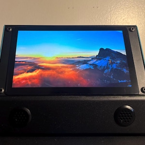 May include: A black and teal electronic device with a screen displaying a mountain landscape at sunset. The device has two circular speaker grills and is mounted with screws.