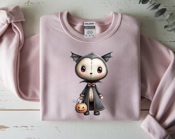 May include: A light pink sweatshirt featuring a cartoon vampire character holding a jack-o'-lantern. The vampire has a gray cape, bat wings, and a bow tie. The sweatshirt is folded neatly on a light-colored wooden surface.