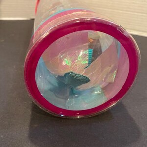 May include: A clear plastic container with a magenta rim and lid, filled with colorful fabric and iridescent materials. The contents include shades of blue, pink, and green, creating a vibrant display. The container is cylindrical.