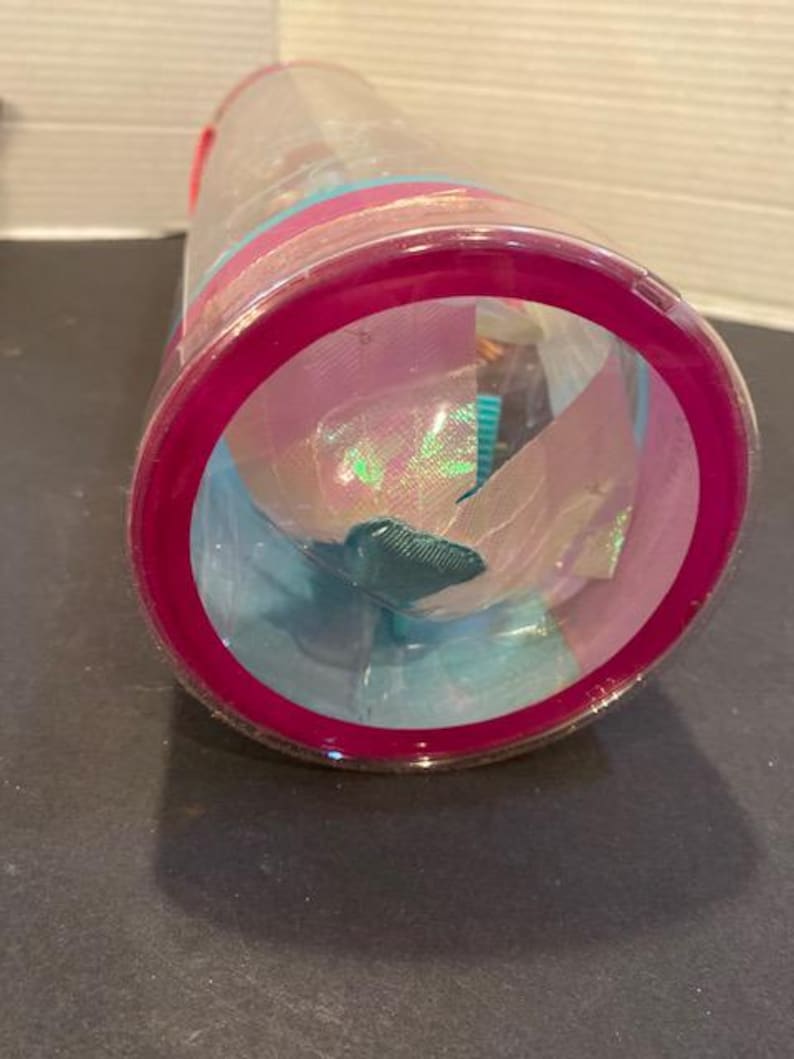 May include: A clear plastic container with a magenta rim and lid, filled with colorful fabric and iridescent materials. The contents include shades of blue, pink, and green, creating a vibrant display. The container is cylindrical.