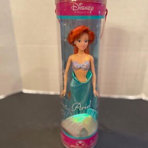 May include: A Disney Princess Ariel doll in a clear plastic tube. The doll has red hair, a silver top, and a teal tail. The tube has pink and teal accents with the Disney Princess logo and the name "Ariel".