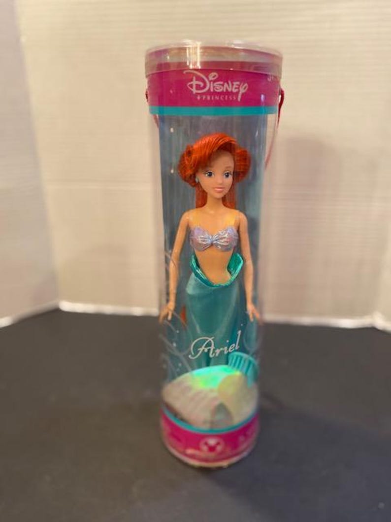 May include: A Disney Princess Ariel doll in a clear plastic tube. The doll has red hair, a silver top, and a teal tail. The tube has pink and teal accents with the Disney Princess logo and the name "Ariel".