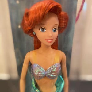 May include: A doll of a red-haired mermaid with blue eyes and a shimmering seashell top. The doll has a teal tail and is a popular character from a children's animated movie.